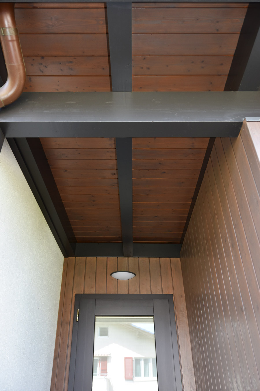 Farbgestaltung Holzfassade: Das Bild zeigt den Eingangsbereich des Atelieranbaus mit der Eingangstür und dem wirkungsvollen Helldunkelkontrast zwischen Holzverschalung und tragenden Balken.